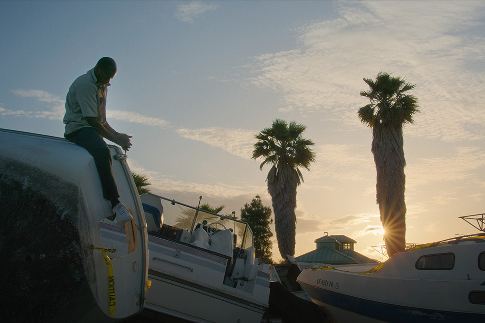A man sitting on a boat with the sun setting behind a palm tree.